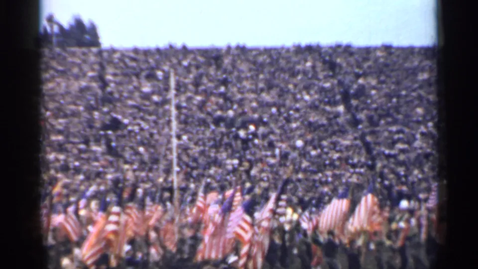 A handheld 8mm view of the San Francisco patriotic ceremony shows flags clustered below a vast audience in the stands.