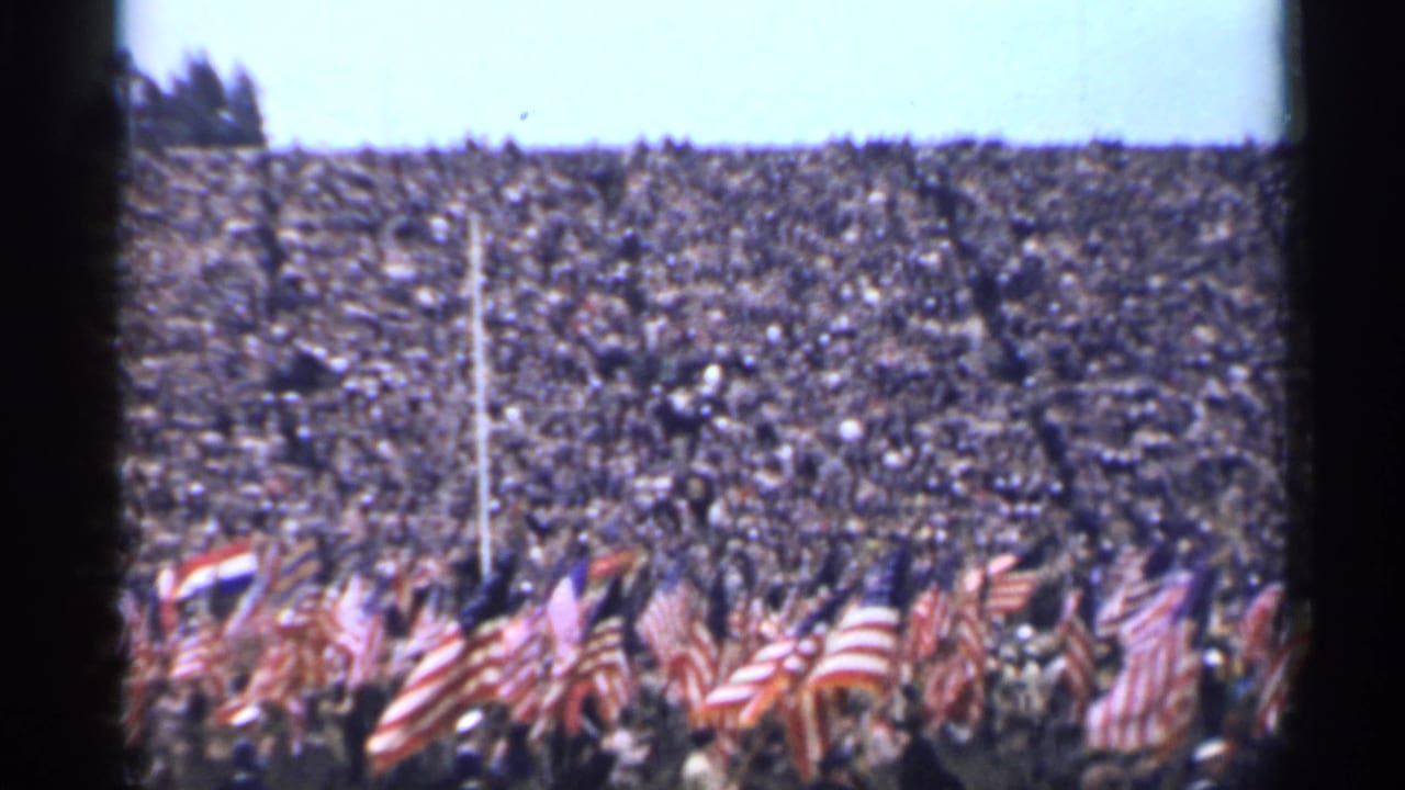 Packed stadium crowd with American flags waving in the foreground.