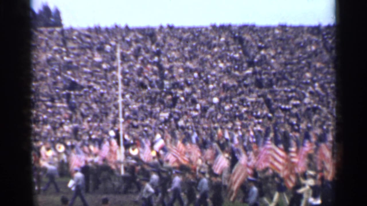 1944 San Francisco home-movie frame showing crowded stadium seating and a field procession with numerous American flags.