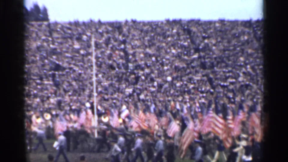 1944 San Francisco home-movie frame showing crowded stadium seating and a field procession with numerous American flags.