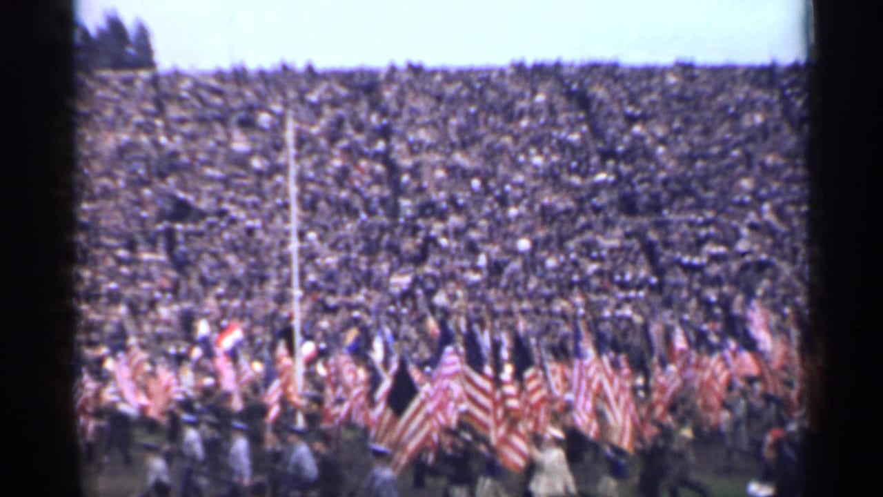 Large crowd in stadium seating as flag bearers move across the foreground.