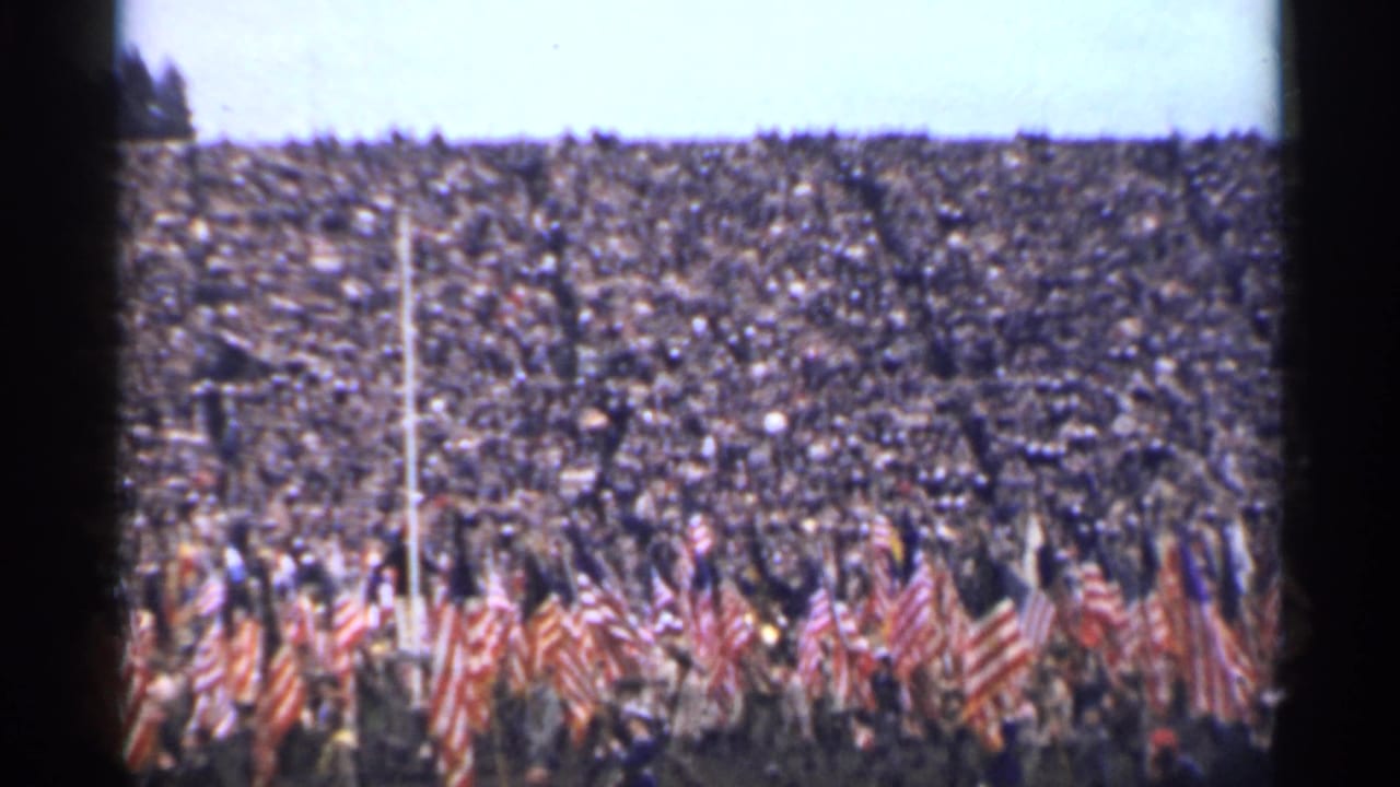 Packed stadium crowd with American flags and marchers in the foreground.