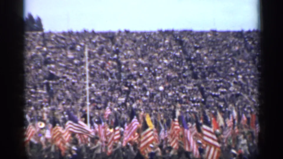 A 1944 San Francisco stadium scene with dense spectators and numerous American flags moving across the lower frame.