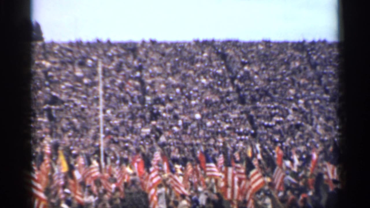 Crowded stadium with American flags raised along the field below.