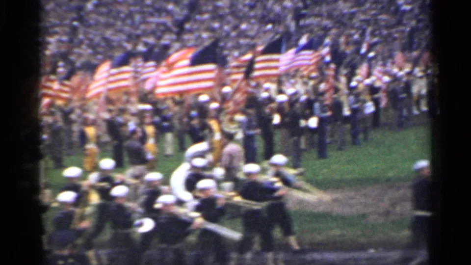1944 San Francisco home-movie frame showing a marching band in dark uniforms and white caps near American flags in a stadium.
