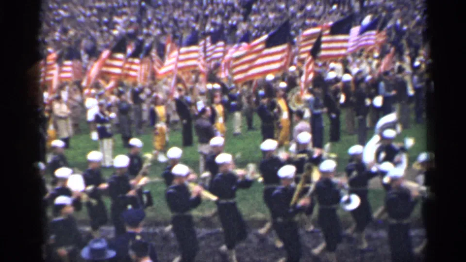1944 San Francisco frame with band members in white caps, American flags, and a packed stadium crowd in the background.