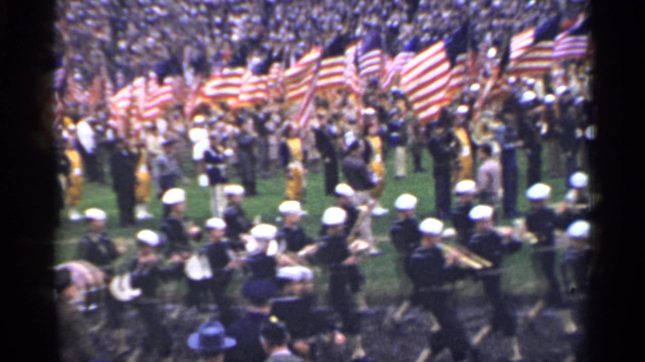 In the supplied 1944 San Francisco home-movie context, uniformed sailors march as a band while spectators stand behind them with many American flags.