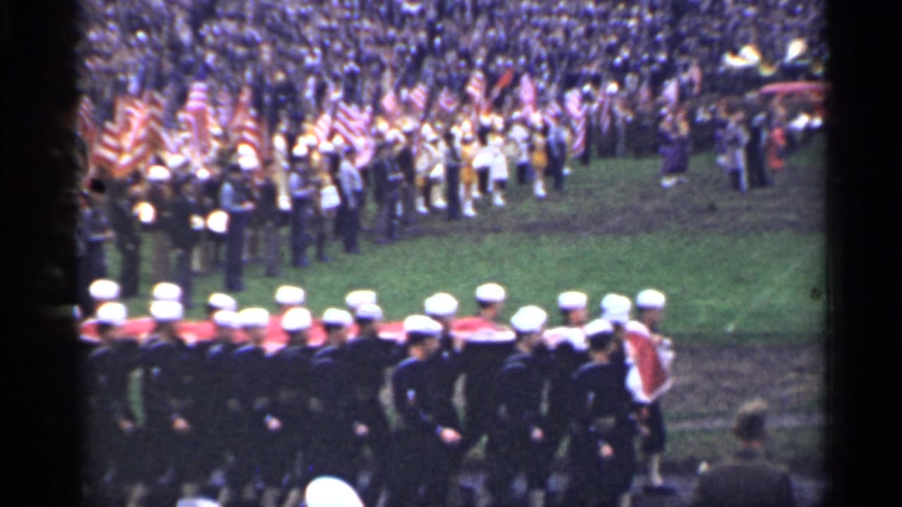 Uniformed sailors with white caps move in formation across the foreground as a packed crowd waves American flags in the stadium setting.