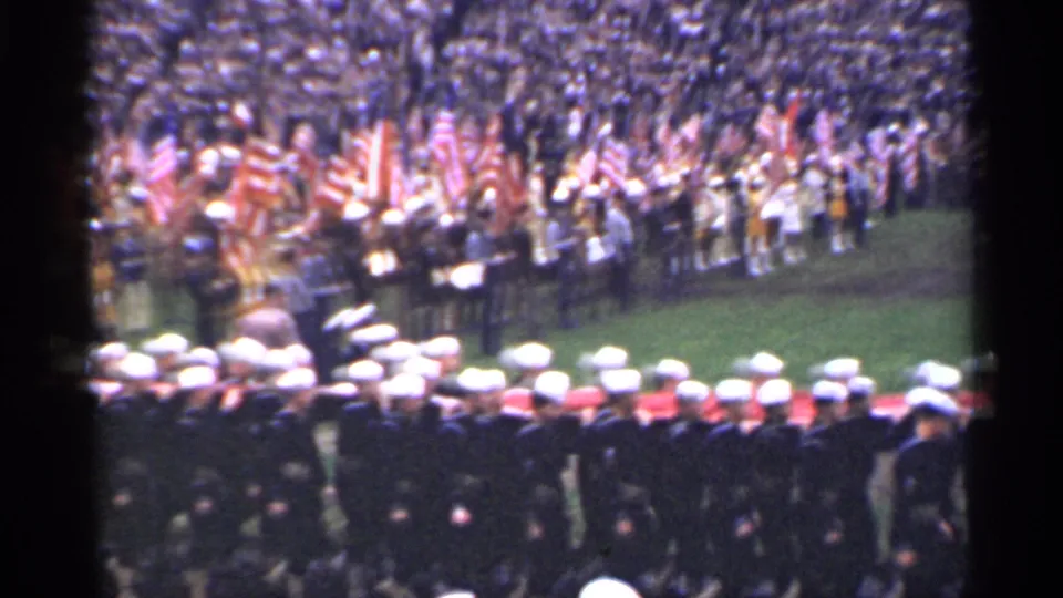 From a spectator vantage point, rows of sailors fill the lower frame while a large flag-bearing crowd stands across the field.