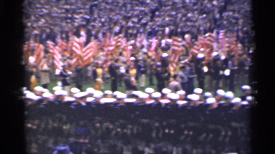 A row of uniformed sailors crosses the foreground while spectators gather with American flags in a crowded 1940s stadium scene supported by the clip metadata.