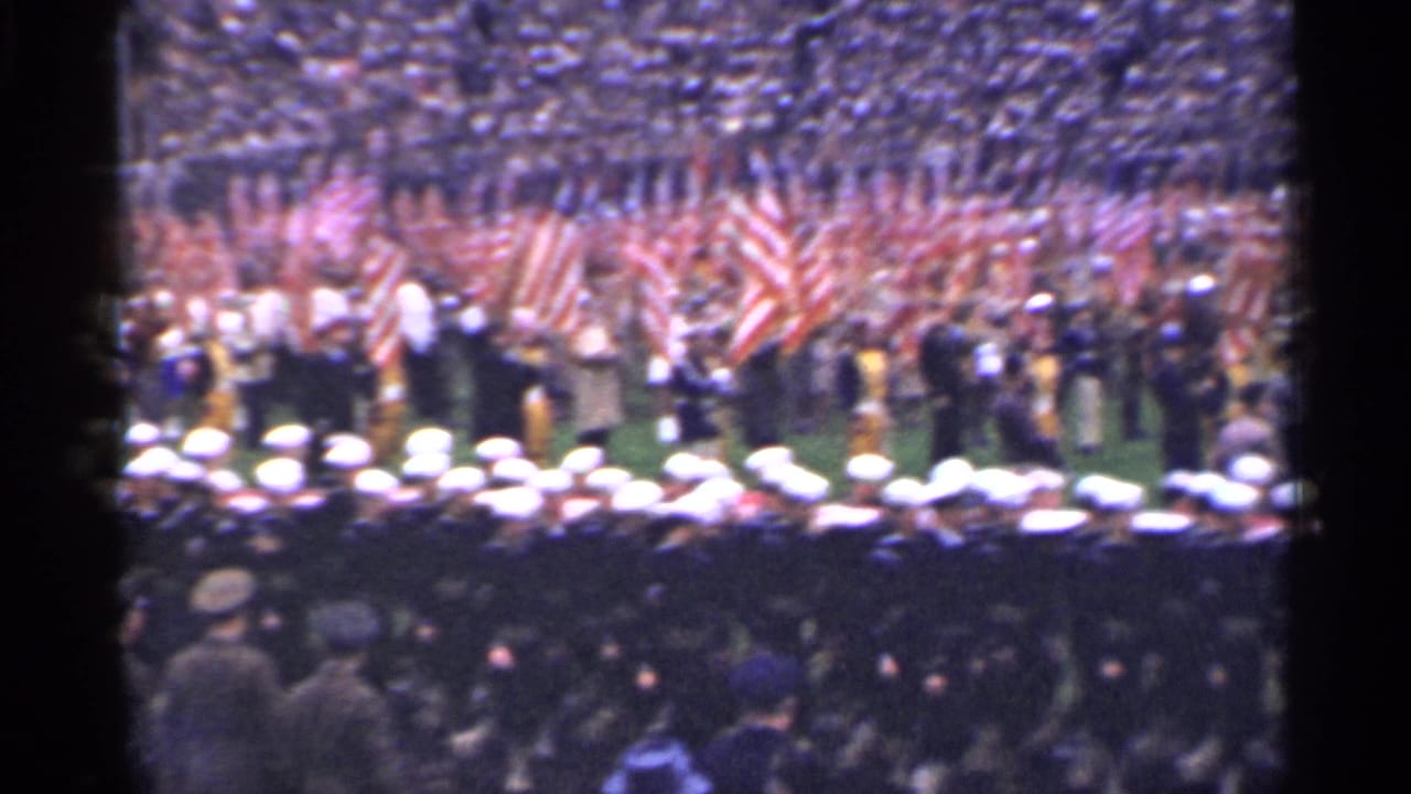 Uniformed personnel face a color guard with American flags before a crowded stadium.