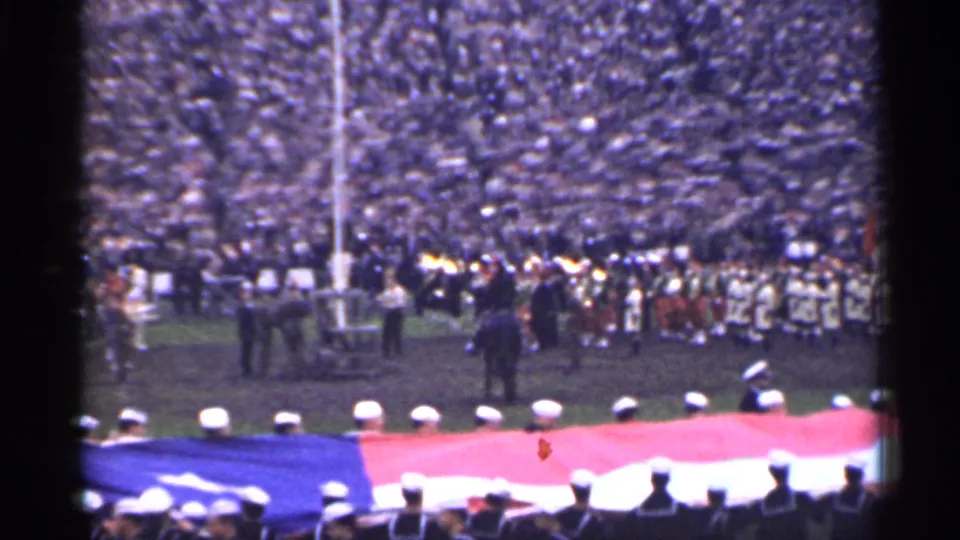 1944 San Francisco ceremony frame with marching band members, uniformed personnel, packed stands, and a large American flag near the camera.