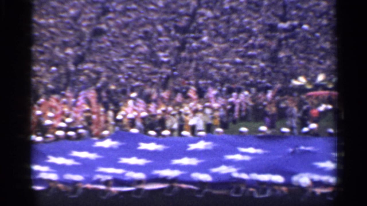 Crowded stadium with uniformed personnel holding the edge of a large American flag.