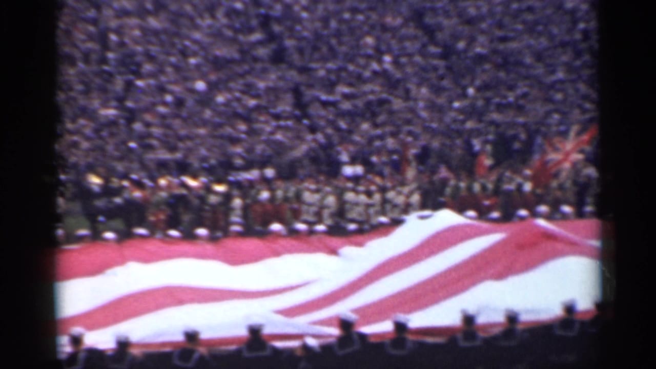 Large American flag spread across a stadium field with crowd-filled bleachers behind it.