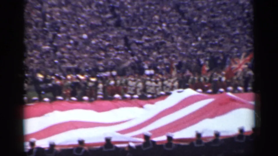 In 1944 San Francisco footage, a giant American flag lies across a stadium field before packed bleachers and uniformed personnel.