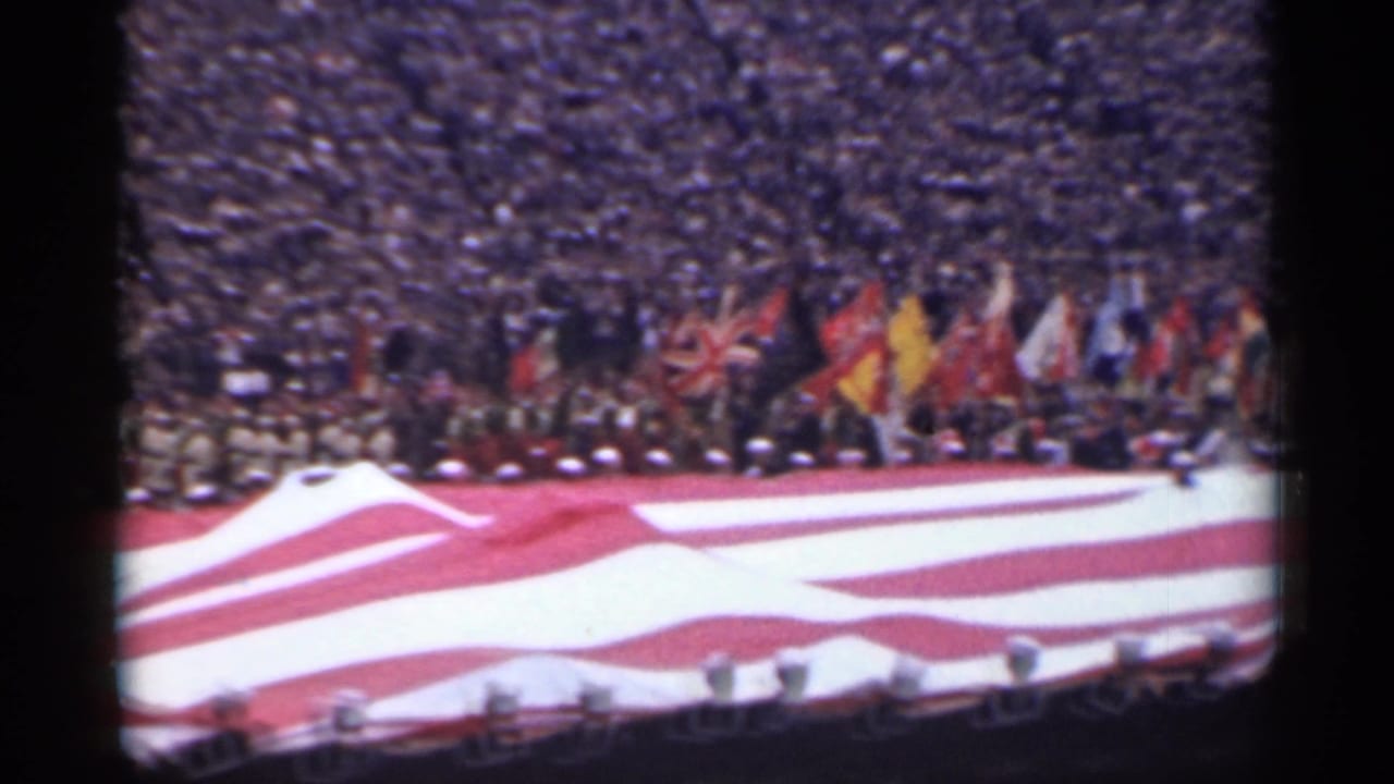American flag stretched across the field with national flags and spectators in the background.