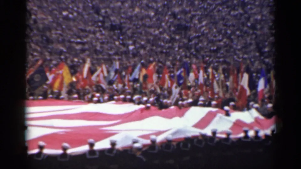 A packed stadium watches a large American flag display with uniformed personnel and many national flags visible in the background.