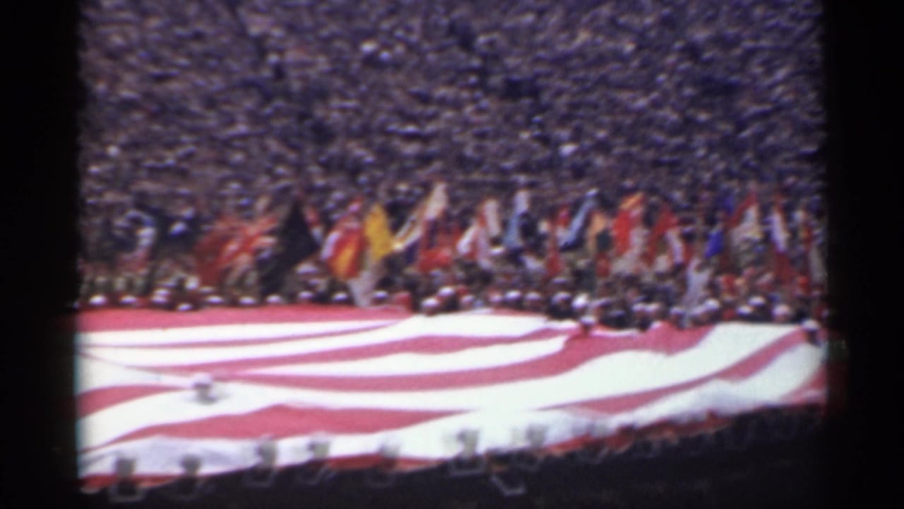 Large American flag spread across a stadium field with a dense crowd in the stands.
