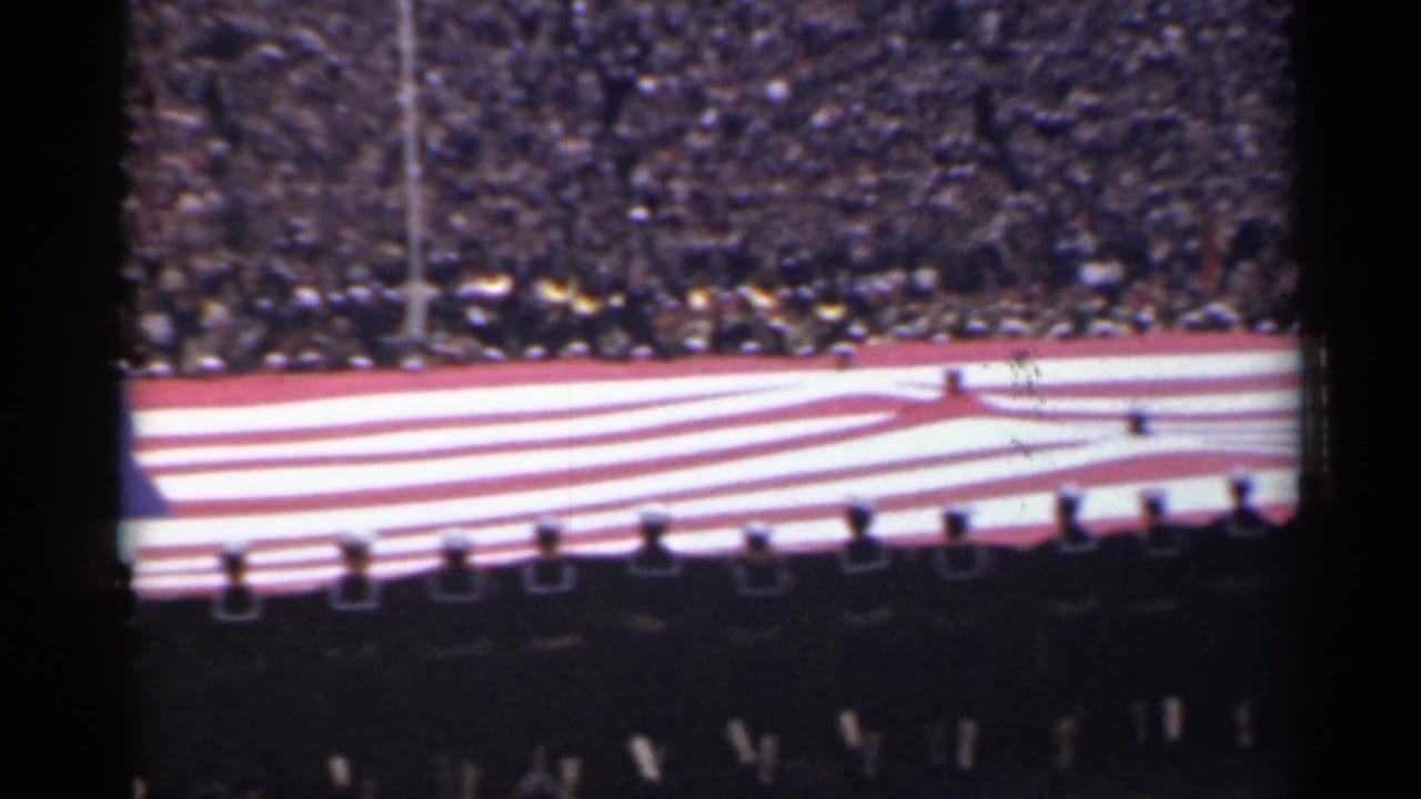 Large U.S. flag carried across a stadium field before packed stands.