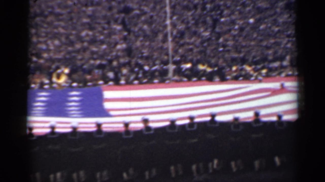 Blue star field of a large U.S. flag passes in front of crowded stands.