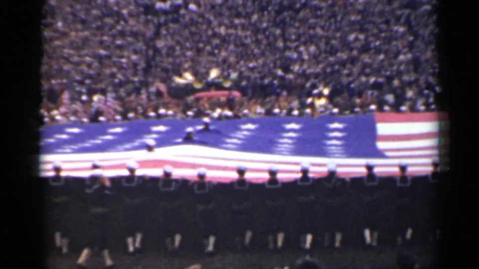 In the stadium, the large American flag stretches across the frame while distant uniformed figures guide it before the audience.