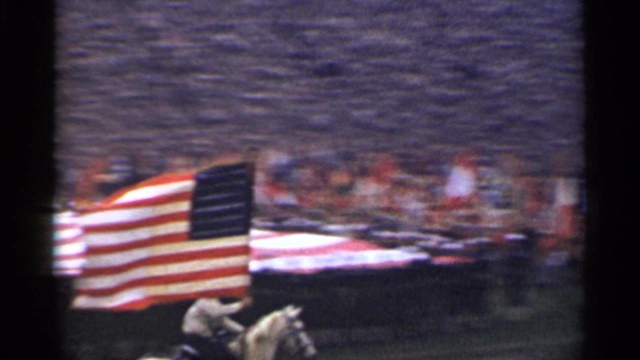 Mounted rider carries a large American flag beside a crowd and field display.