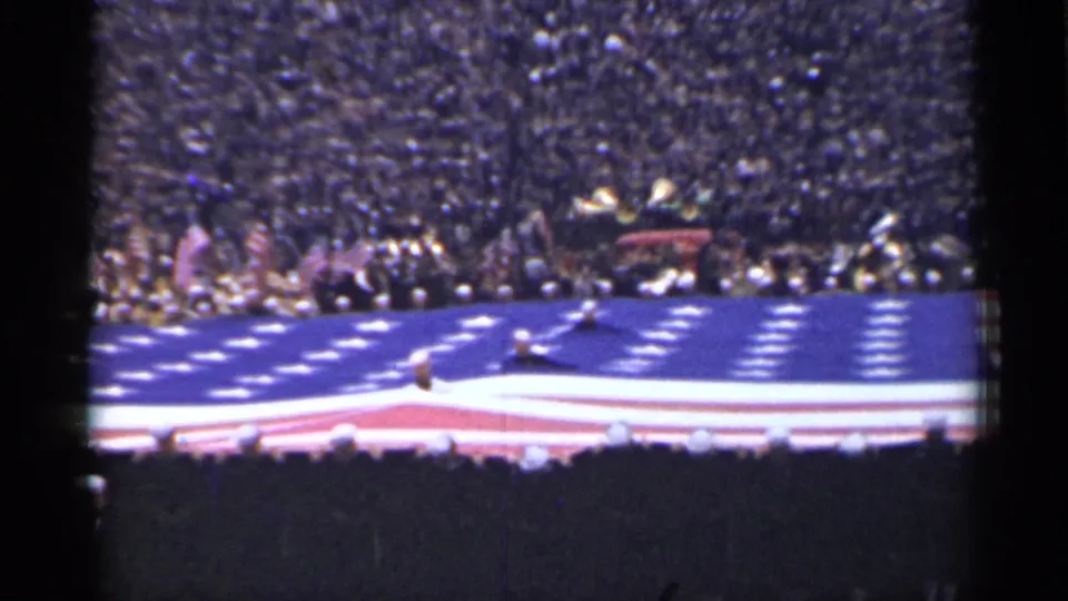 1944 San Francisco frame showing a crowd behind a large U.S. flag spread over an outdoor field.