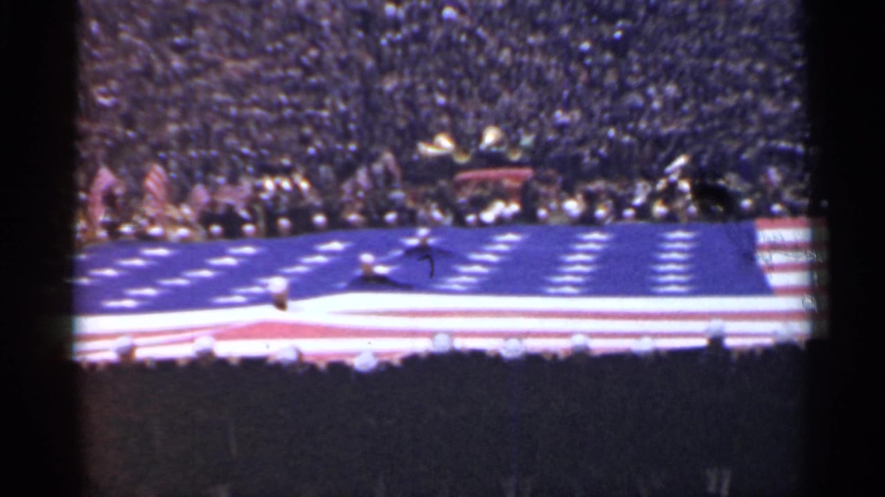Large American flag spread across a football field with players and packed stadium stands behind it.