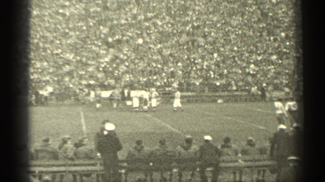 Football players line up on a field before packed stadium stands, with spectators seated and standing near the sideline.