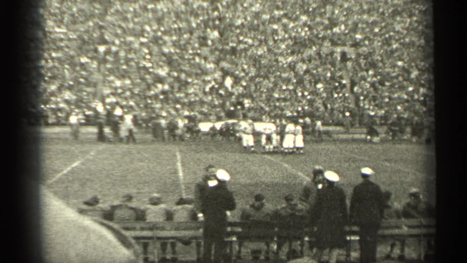 1944 San Francisco football game scene with a player huddle, packed grandstand, sideline benches, and spectators in hats or caps.
