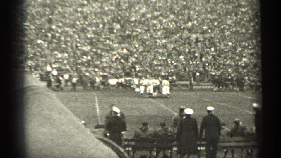 1944 San Francisco football footage showing players near the sideline, dense stadium seating, and foreground spectators by benches.