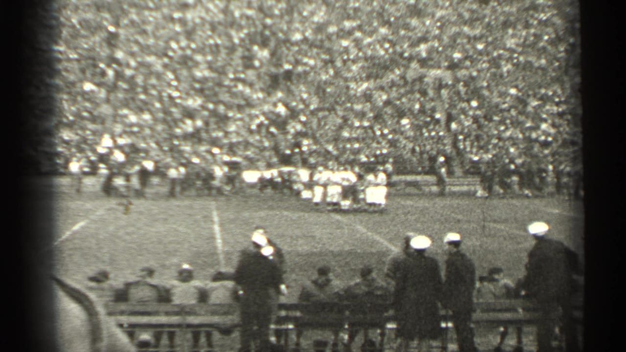 Football game seen from the stands with sideline spectators and players gathered across the field.