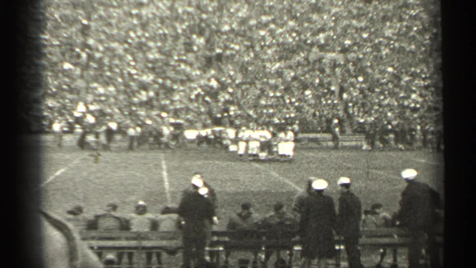 1944 San Francisco 8mm frame showing a football field, crowded stands, sideline benches, and spectators in military-style caps.