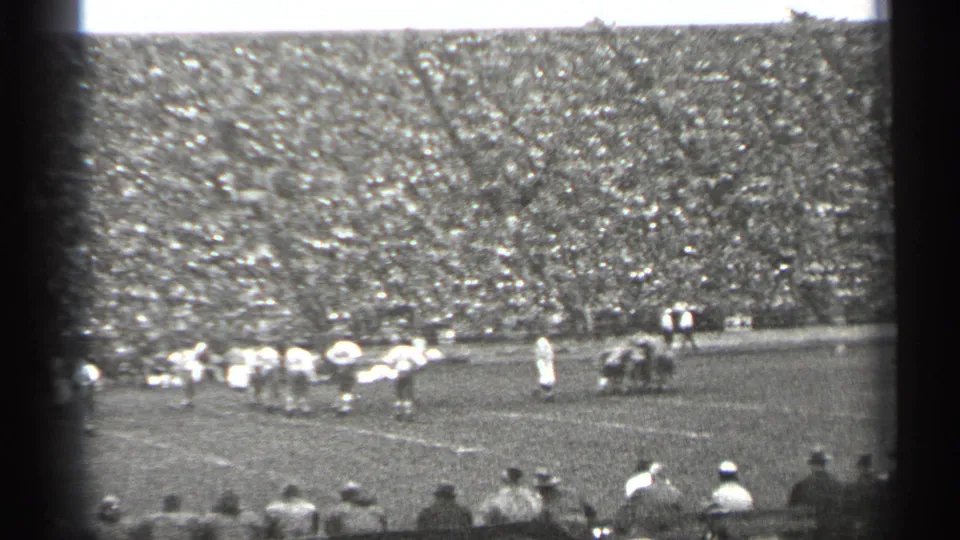 Black-and-white 8mm view of a 1940s San Francisco football huddle, with spectators in the foreground and a crowded stadium behind the field.