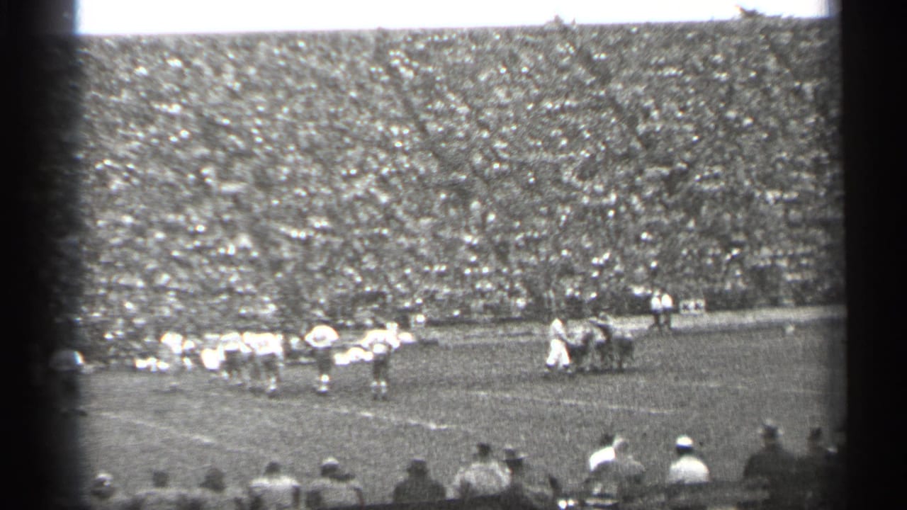 Football players line up on a field before packed stadium stands.
