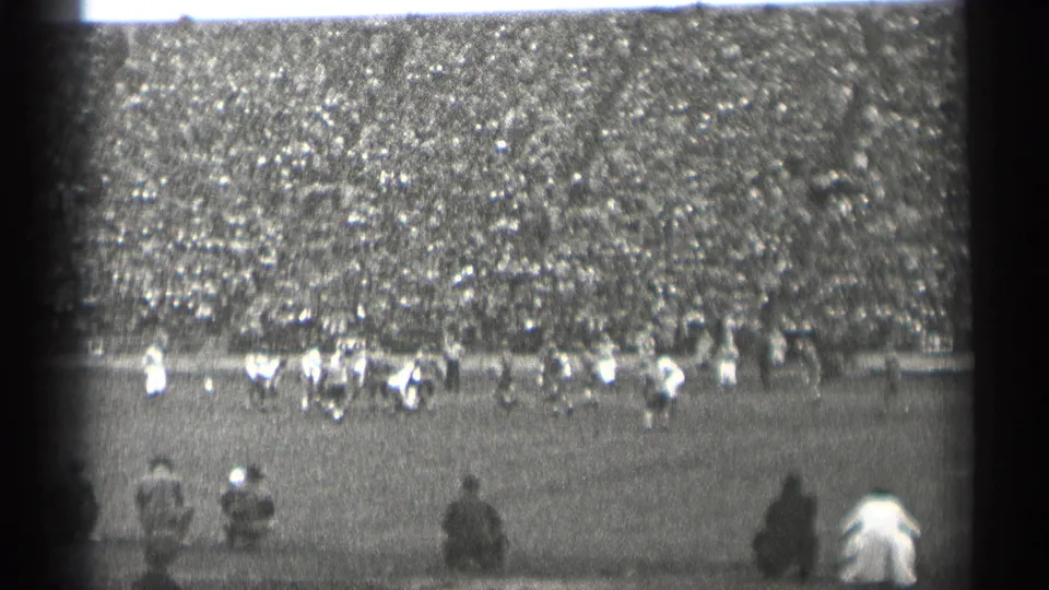 Wide 8mm frame of a 1944 San Francisco football game, with distant players, sideline figures, and a large crowd.