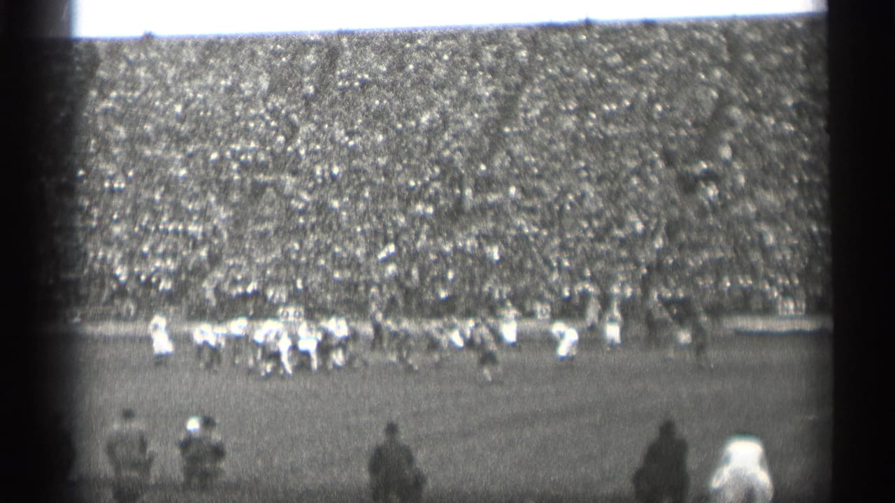Football players run across the field before a crowded grandstand.