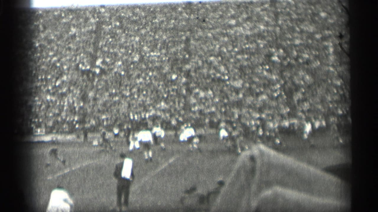 1944 San Francisco football scene viewed from the stands, with players on the field, a sideline figure in a light shirt, and packed spectators beyond.