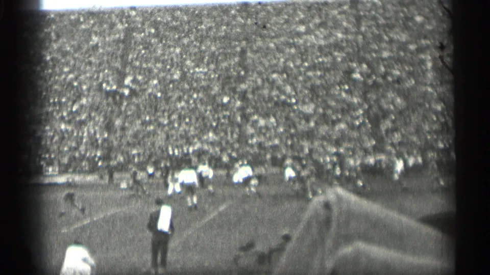 1944 San Francisco football scene viewed from the stands, with players on the field, a sideline figure in a light shirt, and packed spectators beyond.