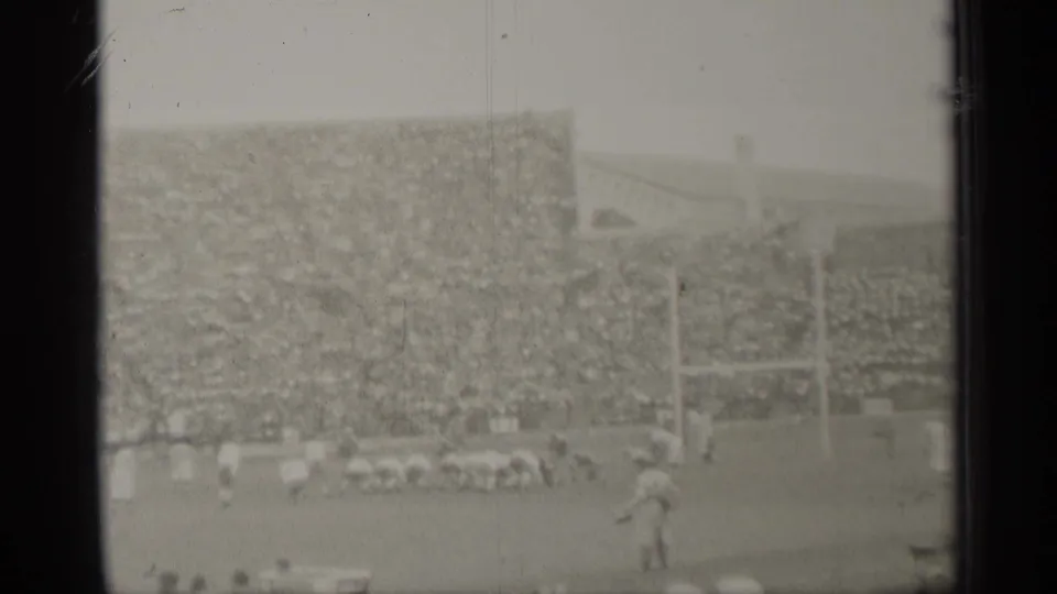 Soft 8mm view of a 1944 San Francisco football game, with players near the line, goal posts, sideline figures, and densely filled stands.