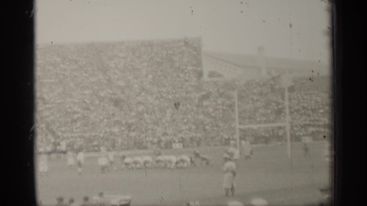 Football players on a field below packed stadium stands and goalposts.