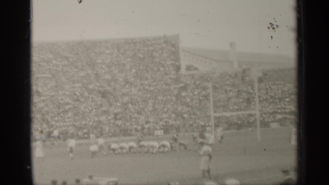 Wide stadium view with football players, goalposts, and crowded stands.