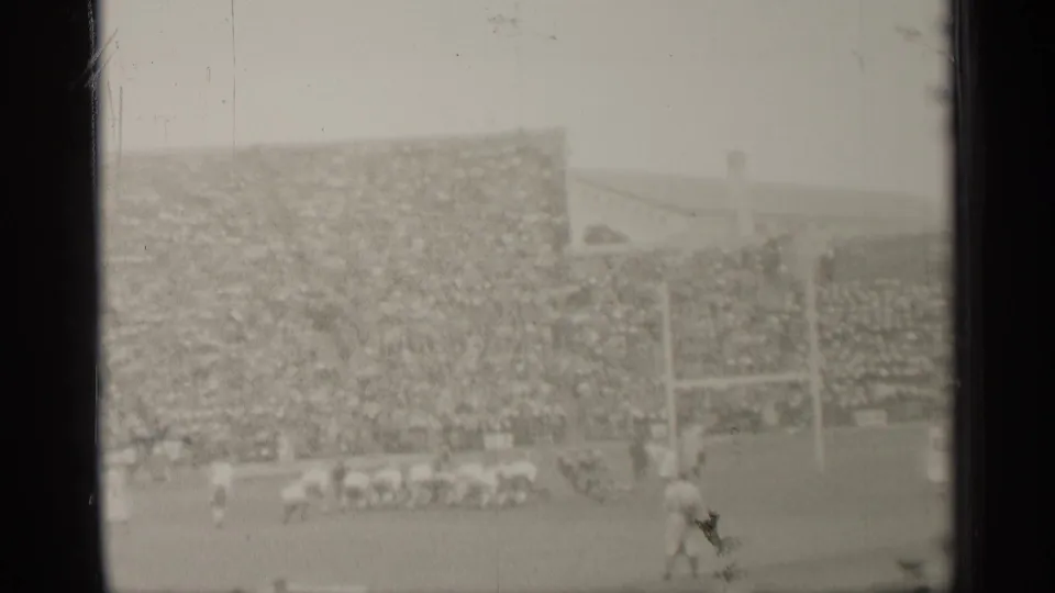 Distant 1944 football frame showing a foreground player, clustered sideline figures, goalposts, and packed San Francisco stadium seating.