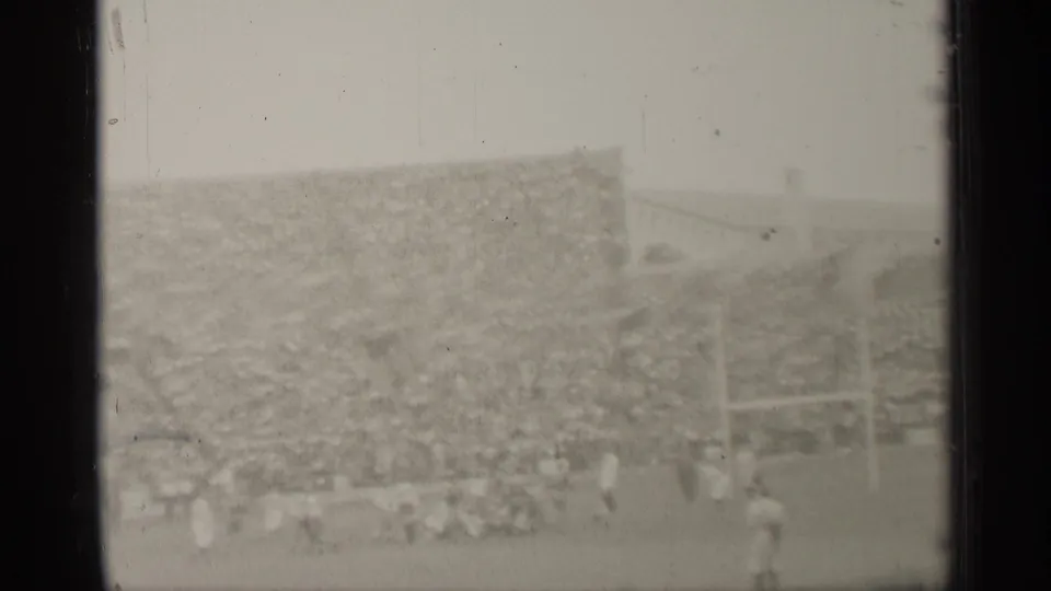 Soft, grainy 8mm view of the San Francisco football game with goalposts, players, sideline figures, and a full stadium crowd.