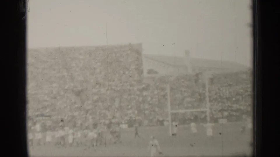 Distant 8mm view of football players crossing the field near goalposts with San Francisco stadium seating behind them.