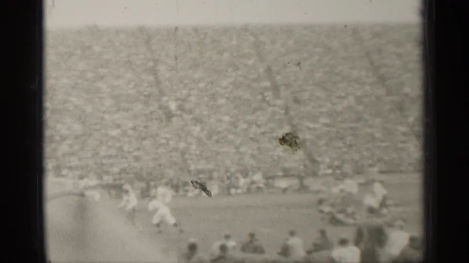 Black-and-white 8mm frame from a San Francisco football game, showing distant field action and a dense crowd in the stands.