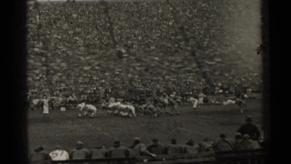 Black-and-white 8mm view of football players on the field with sideline spectators and a crowded San Francisco stadium backdrop.