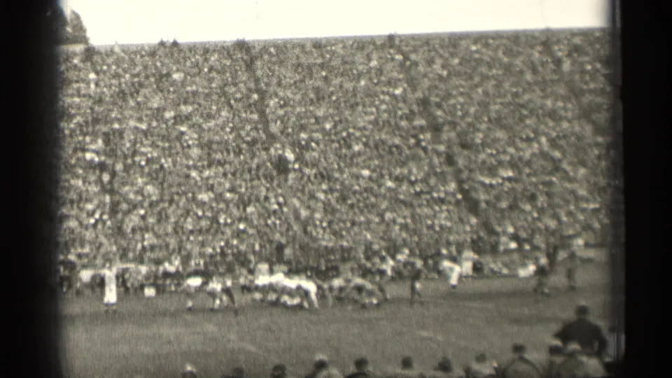 Wide black-and-white 8mm frame of a 1940s football game, with players set on the field and a packed San Francisco crowd behind them.