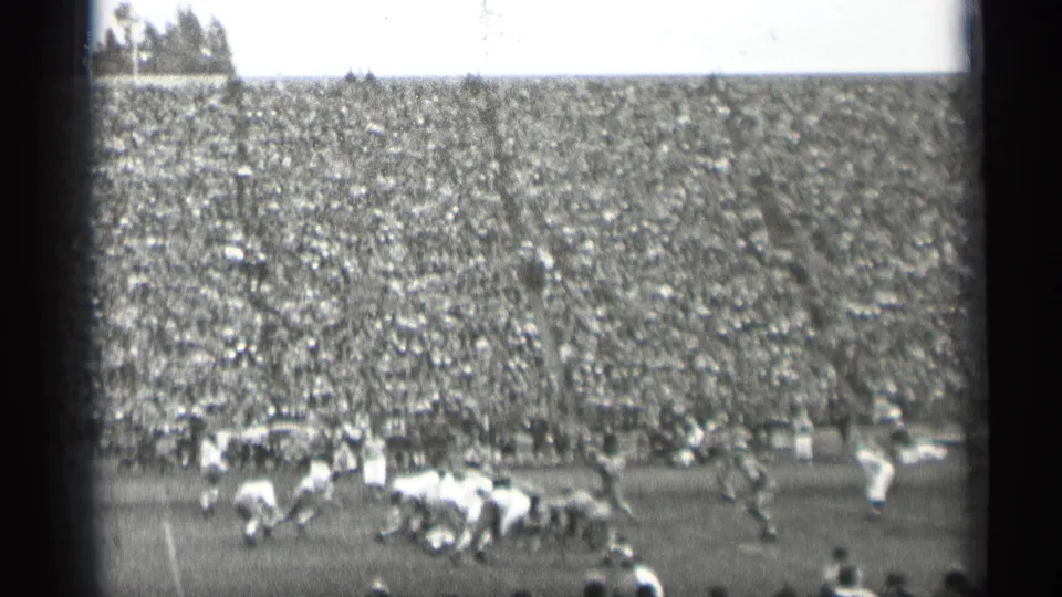 A 1944 San Francisco football play develops near the line, framed by a full stadium crowd and soft black-and-white 8mm texture.