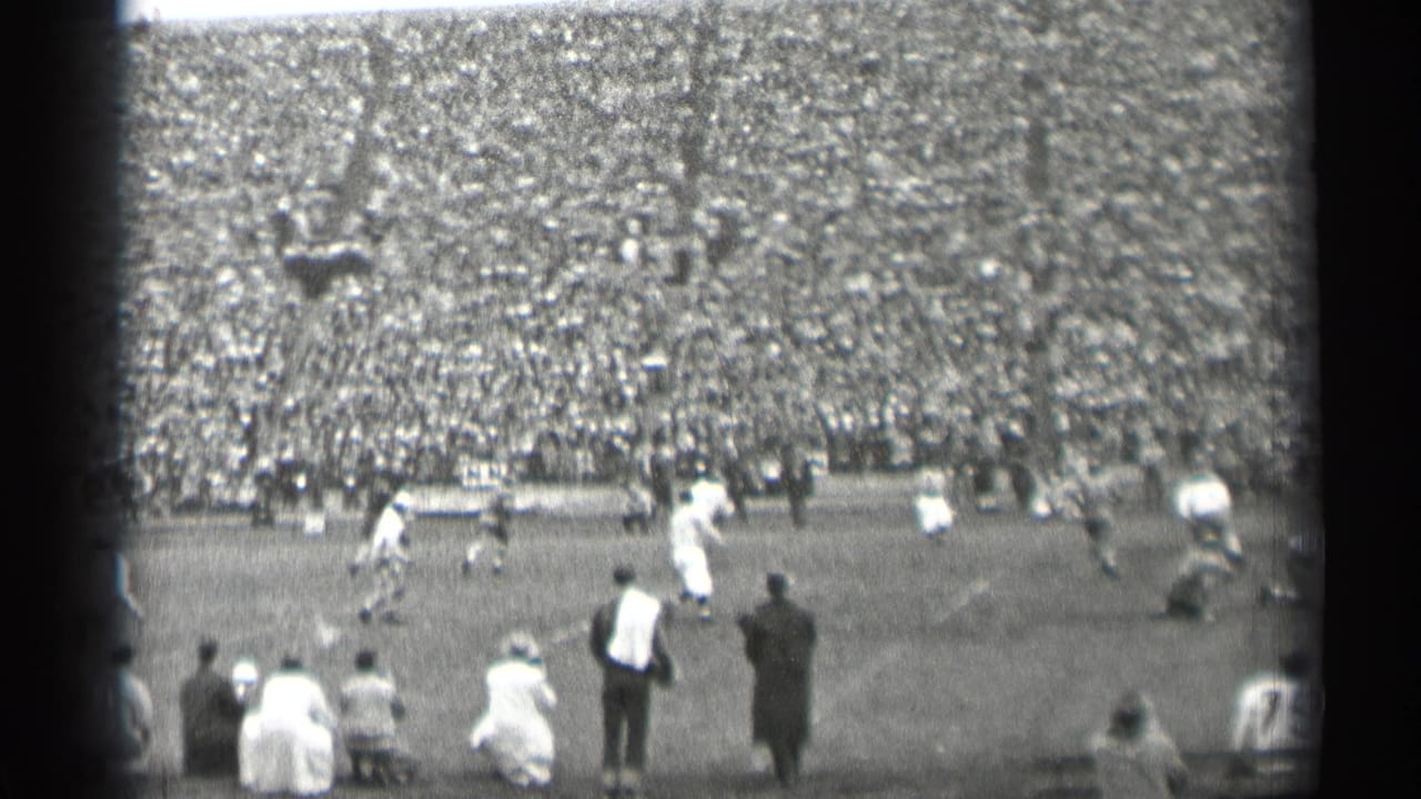 Football play seen from the sideline with a large crowd in the stands.